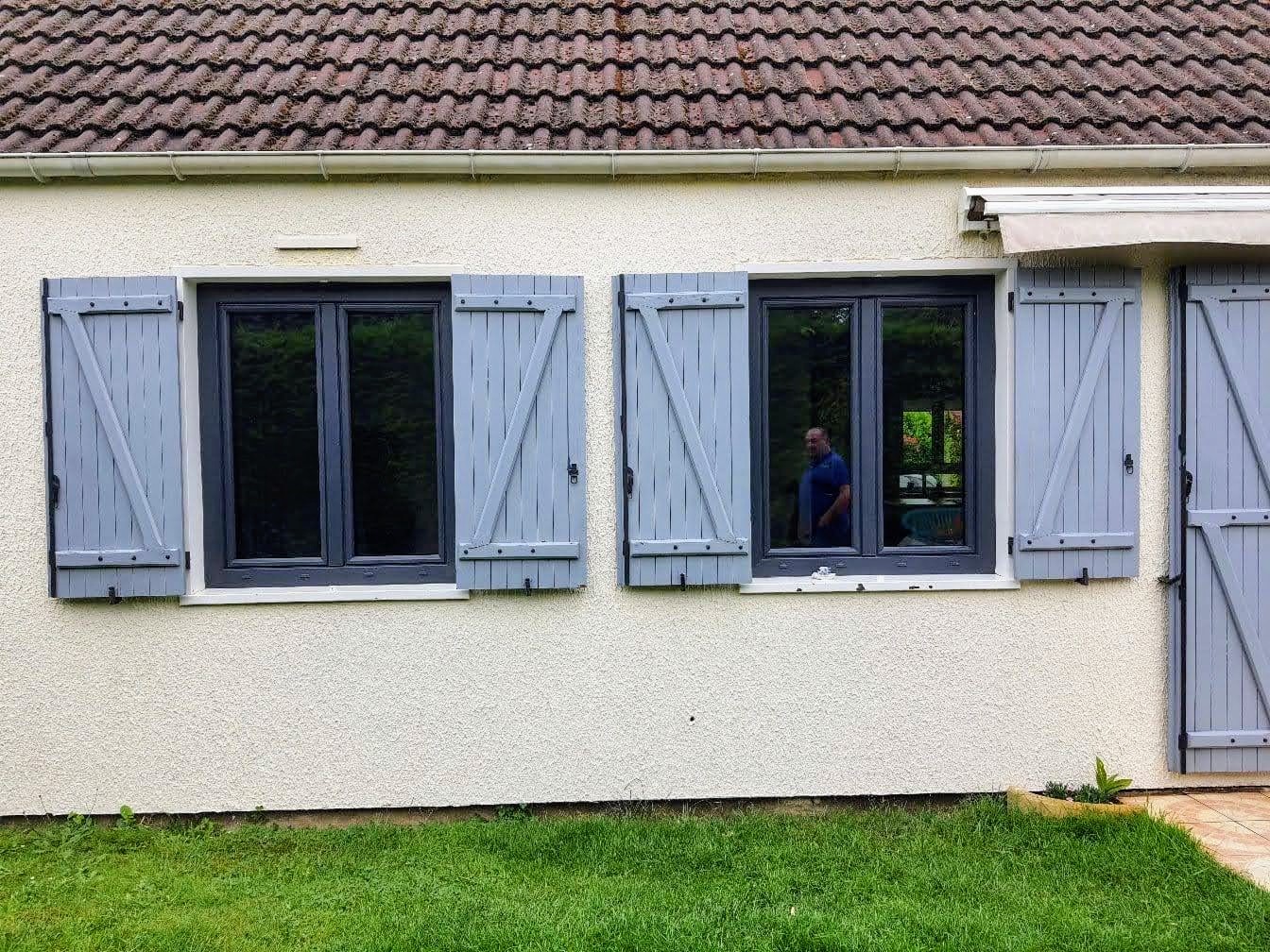 Façade enduite claire avec deux fenêtres neuves en aluminium anthracite, volets battants bois gris-bleu conservés, un poseur visible à l'intérieur