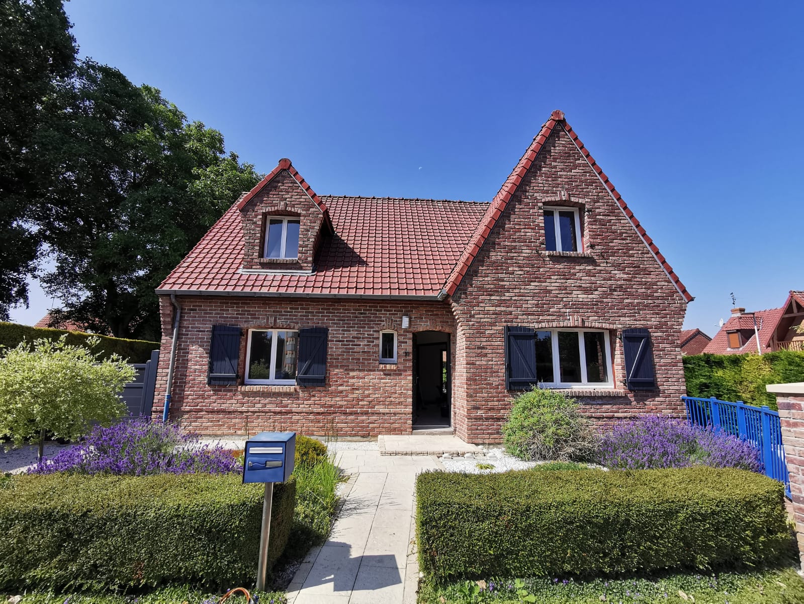 Maison de brique à deux pignons avec fenêtres blanches, volets bleu nuit et jardin entretenu, dans le Nord