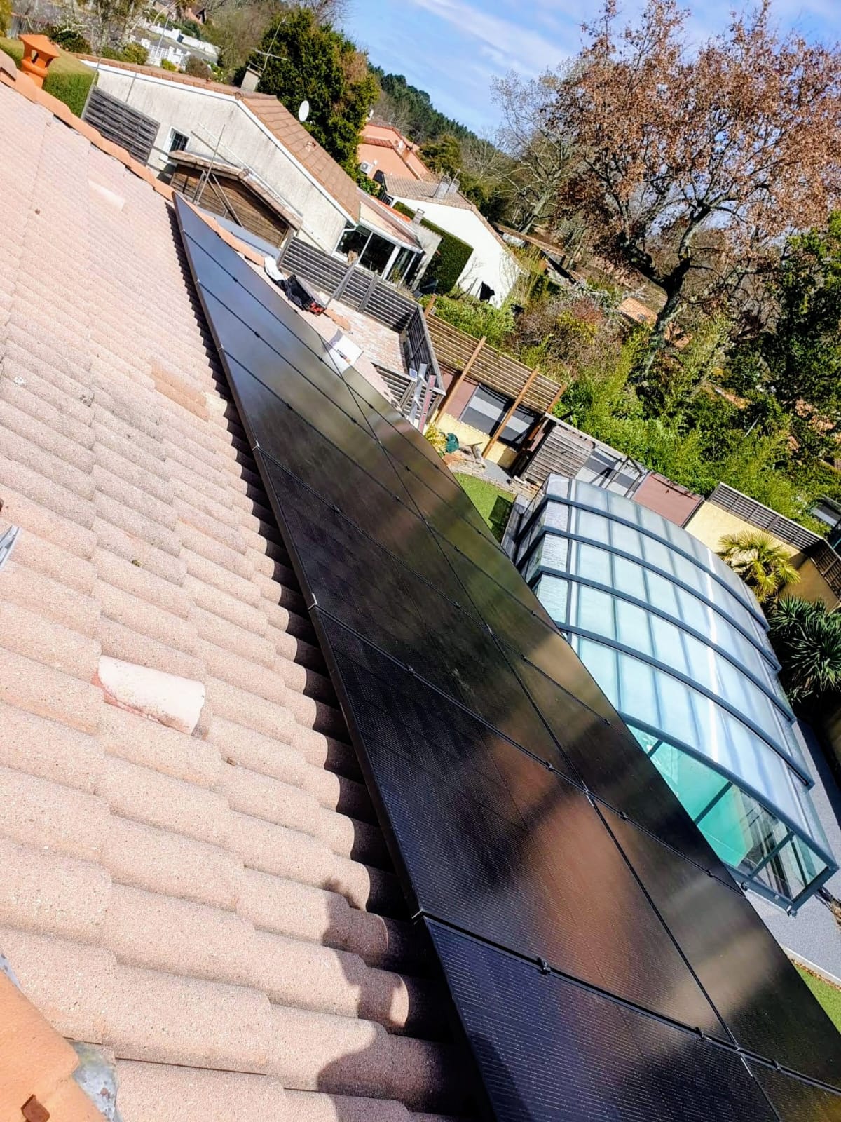 Rangée de panneaux photovoltaïques en surimposition sur une toiture en tuiles, abri de piscine et coteau boisé en arrière-plan