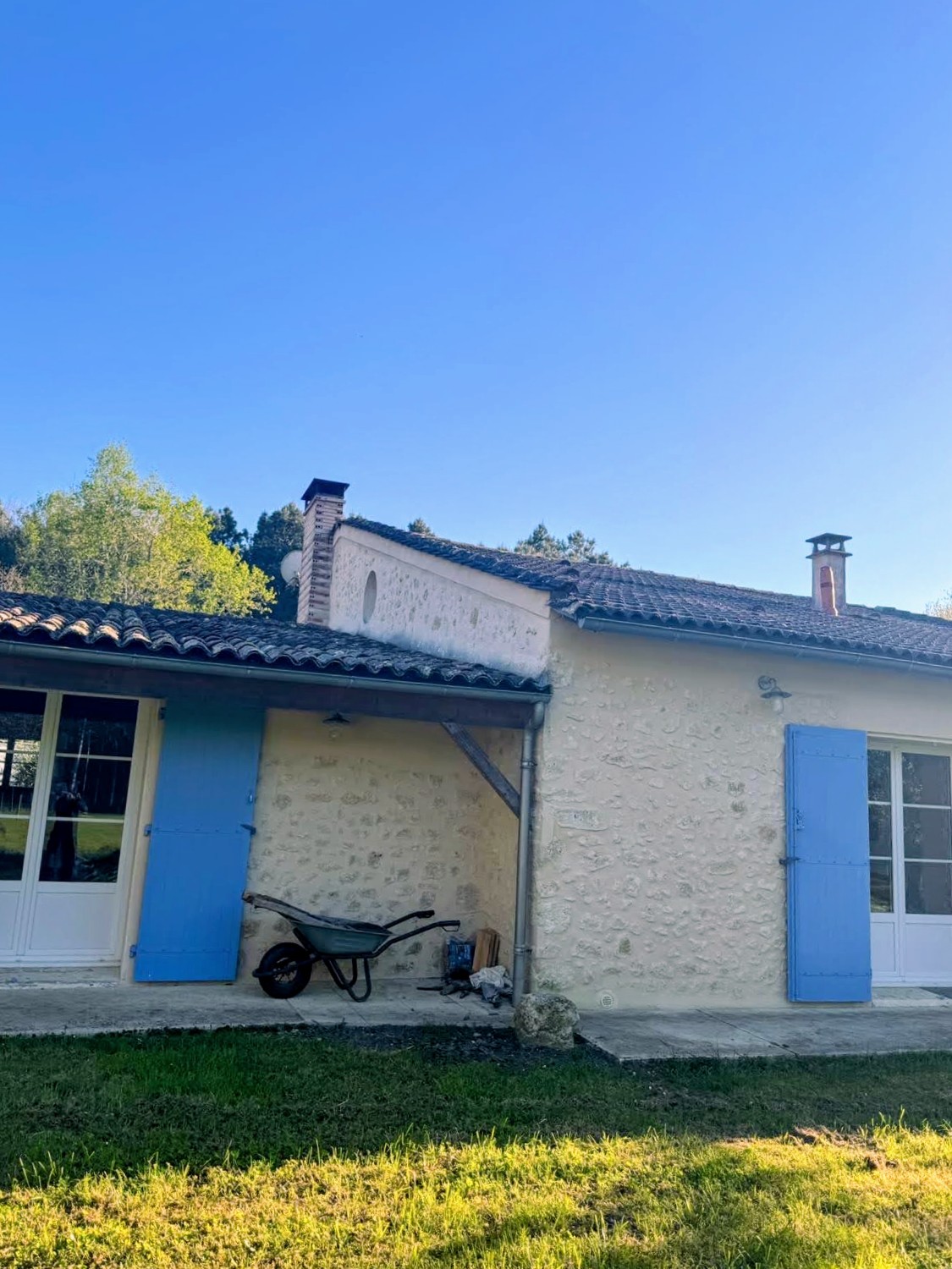 Arrière d'une maison en pierre avec volets bleus, auvent, brouette et matériaux posés au sol