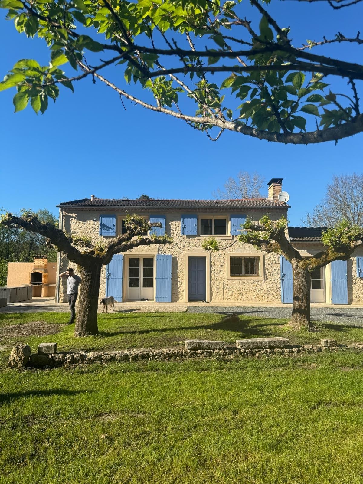 Maison en pierre à deux niveaux avec volets bois bleu clair, arbres taillés au premier plan, une personne adossée à un tronc