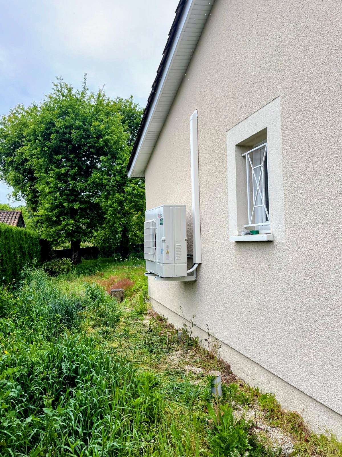 Unité extérieure Mitsubishi Electric fixée en pignon, goulotte blanche alignée le long du mur, jardin tondu au premier plan